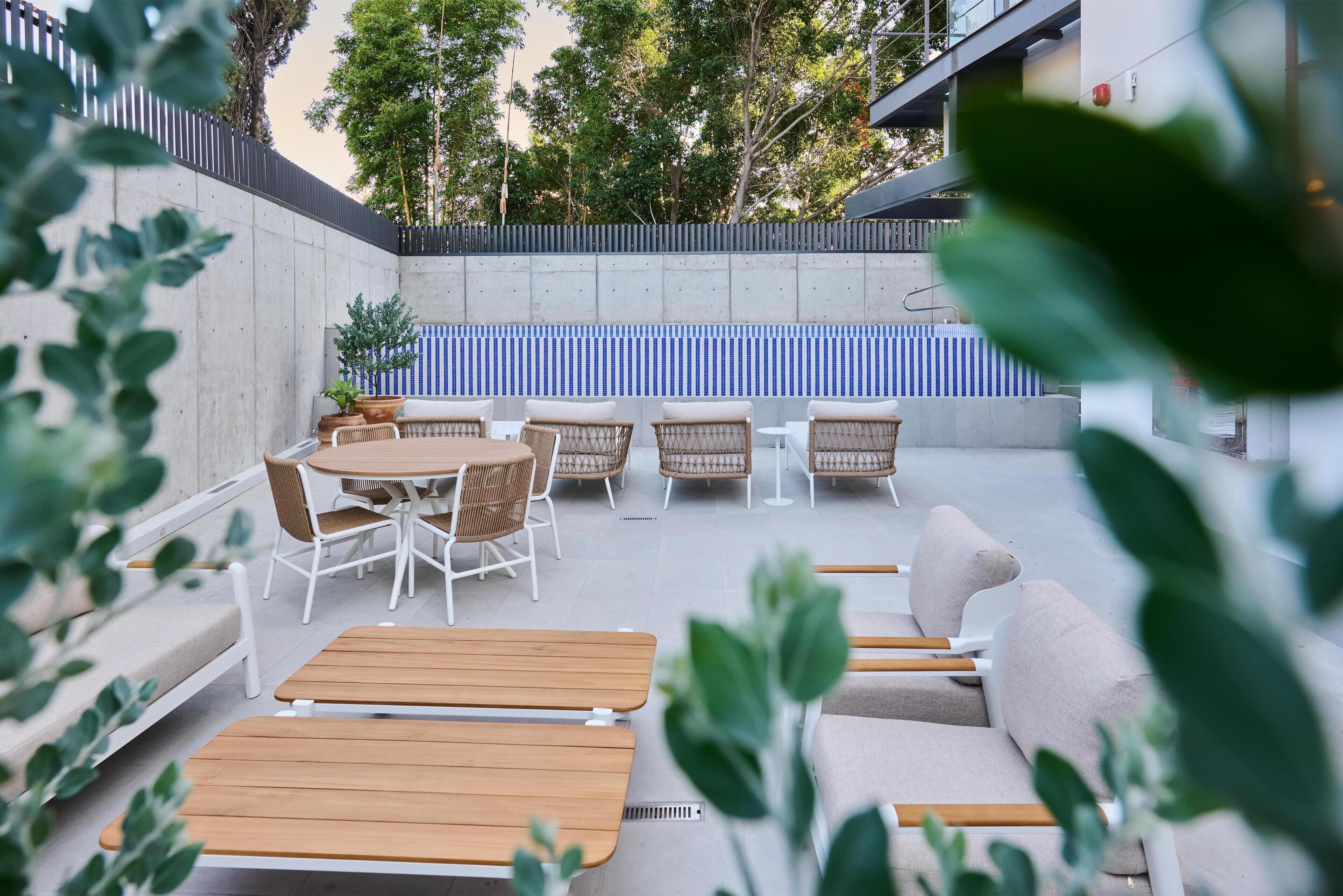 An outdoor seating area with wooden tables and wicker chairs, a concrete wall with a blue and white striped pattern, and a potted plant.