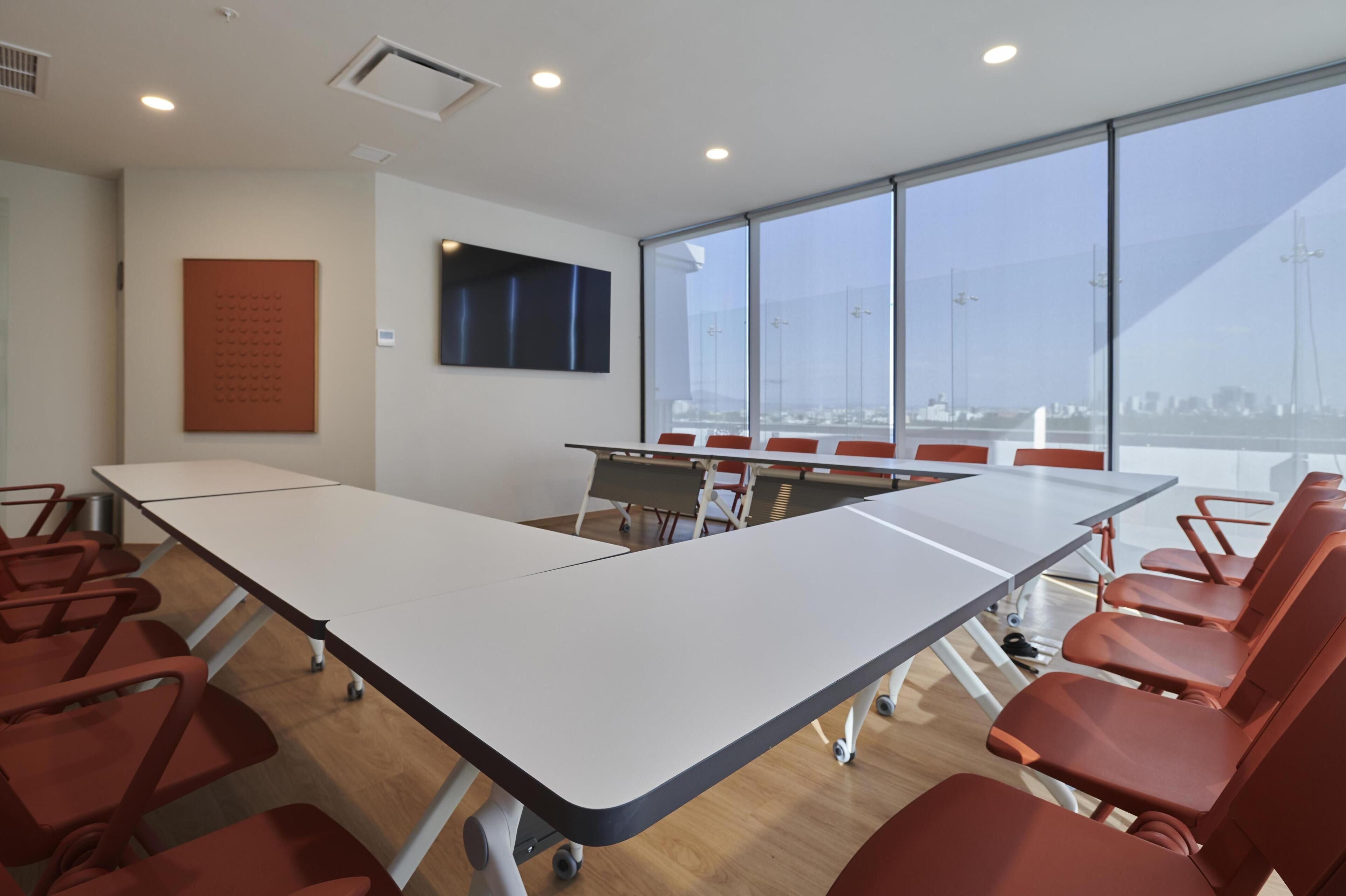 Un salón de conferencias con una larga mesa, sillas rojas, un televisor montado y ventanas amplias con vista a la ciudad.