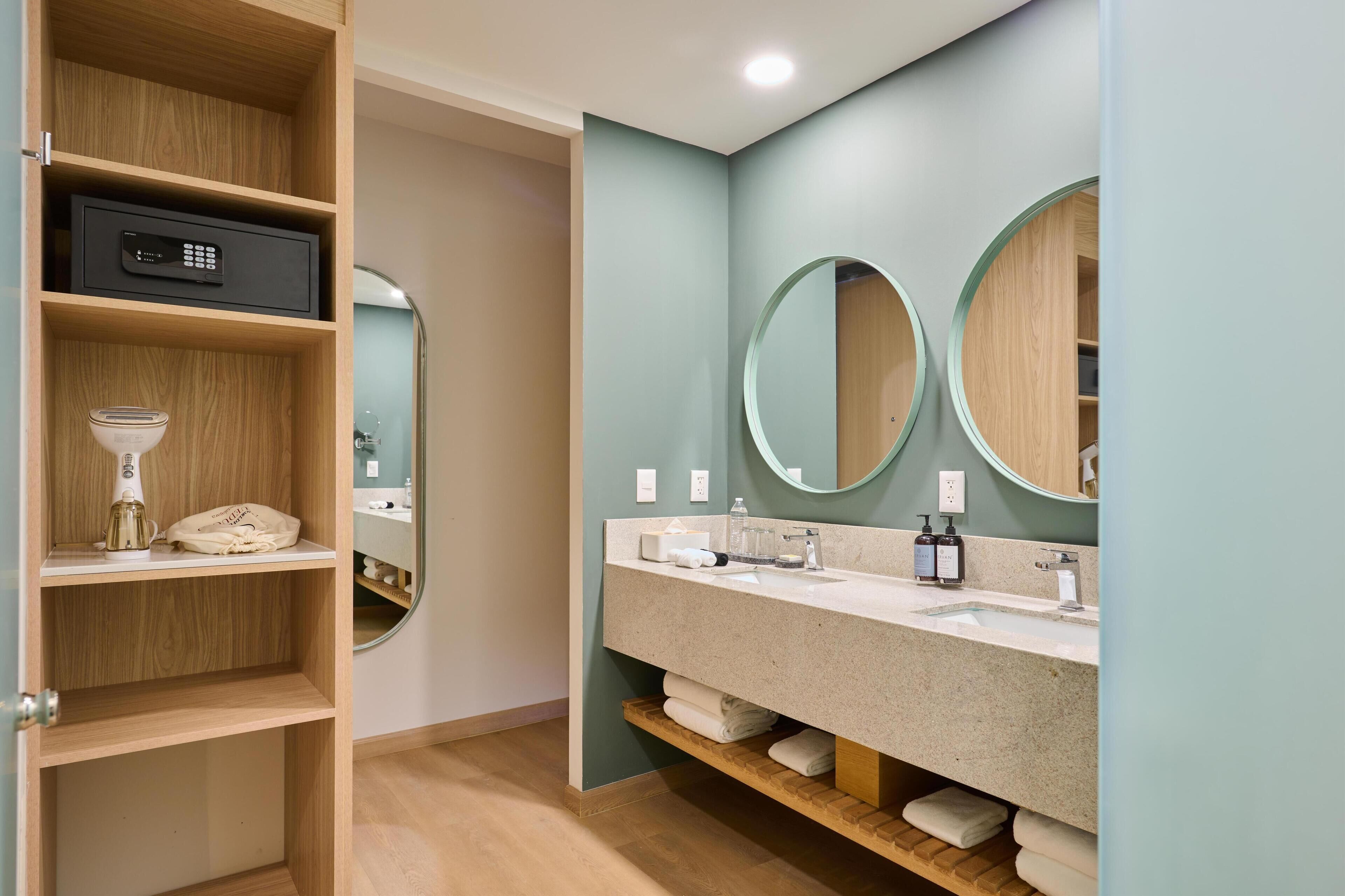 A modern bathroom with a wooden vanity, two round mirrors, a built-in heater, and a shelf with towels.