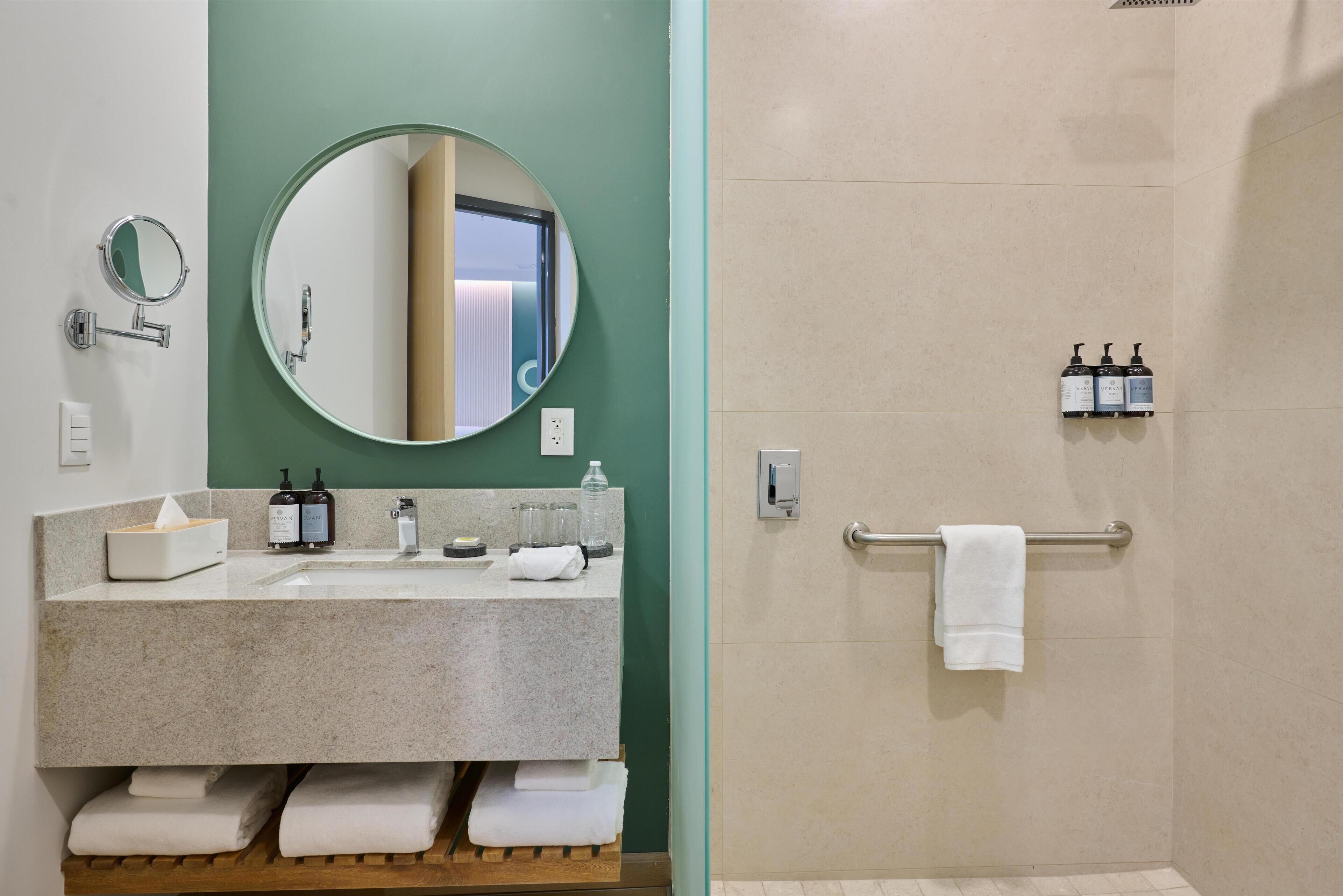 A bathroom with a granite sink, a round mirror, a towel rack, and a shower area.