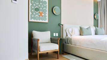 A bedroom with a bed, a chair, a bedside table, a wall-mounted light, and a framed artwork.