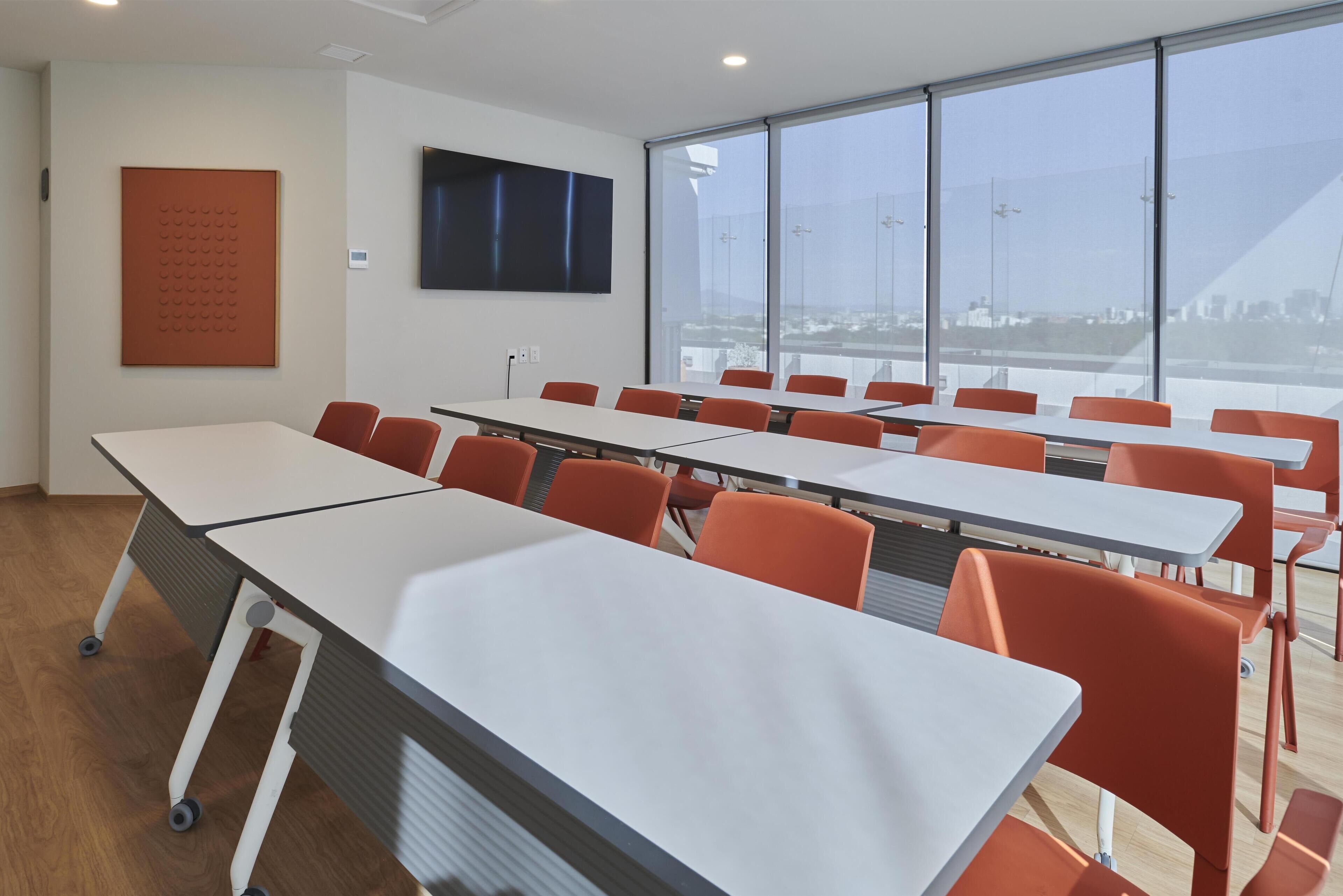 A conference room with white tables and orange chairs, large windows with a city view, a mounted TV, and a poster on the wall.
