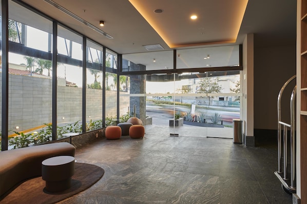 A modern lobby with large windows, a seating area with round ottomans, and a view of the street outside. - WorldHotels Guadalajara Zona Andares (Zapopan)