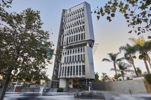 Un edificio moderno de varios pisos con una entrada central, rodeado de palmeras y una fuente.