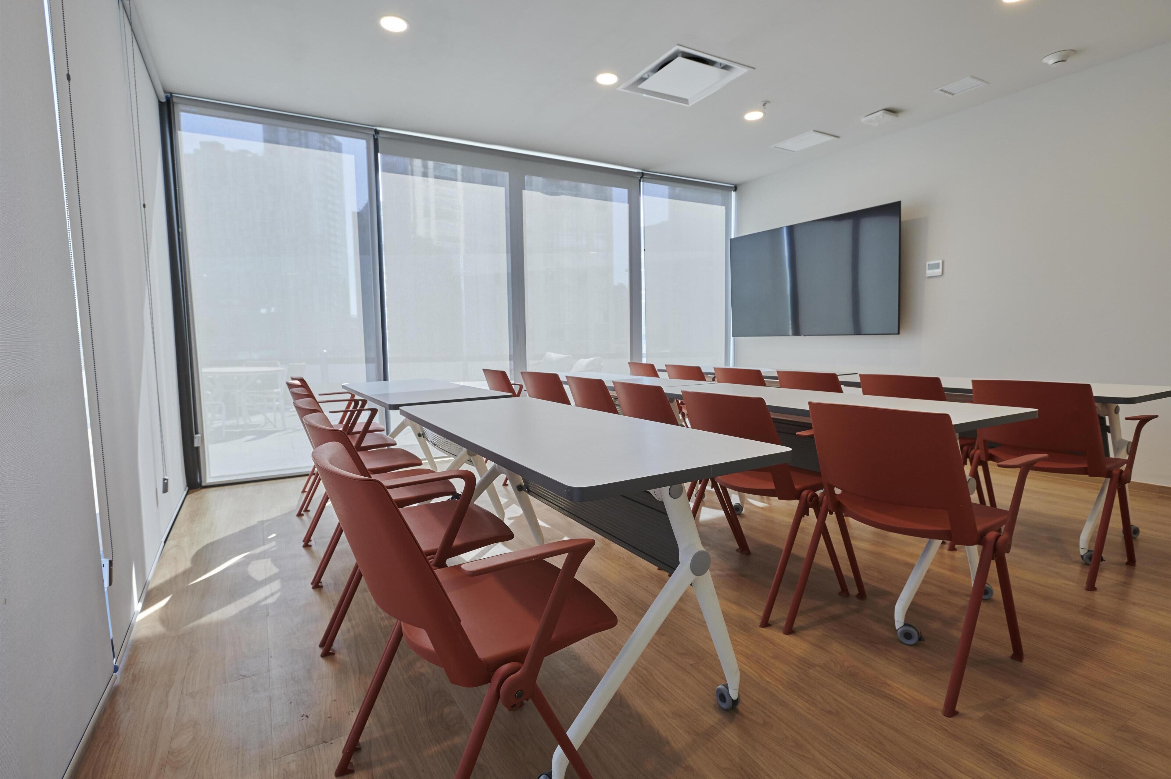 A conference room with a long table and red chairs, large windows, and a flat-screen TV on the wall.