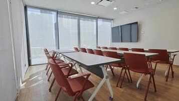 A conference room with a long table and red chairs, large windows, and a flat-screen TV on the wall.