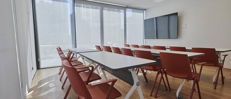 A conference room with a long table and red chairs, large windows, and a flat-screen TV on the wall.