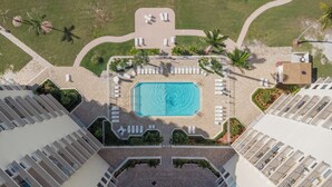 Luftaufnahme eines Hotels mit einem zentral gelegenen Schwimmbad, umgeben von Liegestühlen und Palmen.