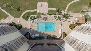 Luftaufnahme eines Hotels mit einem zentral gelegenen Schwimmbad, umgeben von Liegestühlen und Palmen.