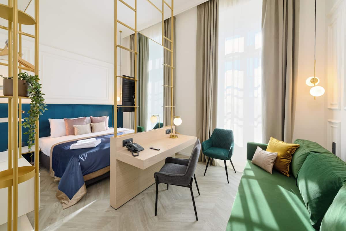A hotel room with a bed, desk, chair, green sofa, and a large window with curtains.