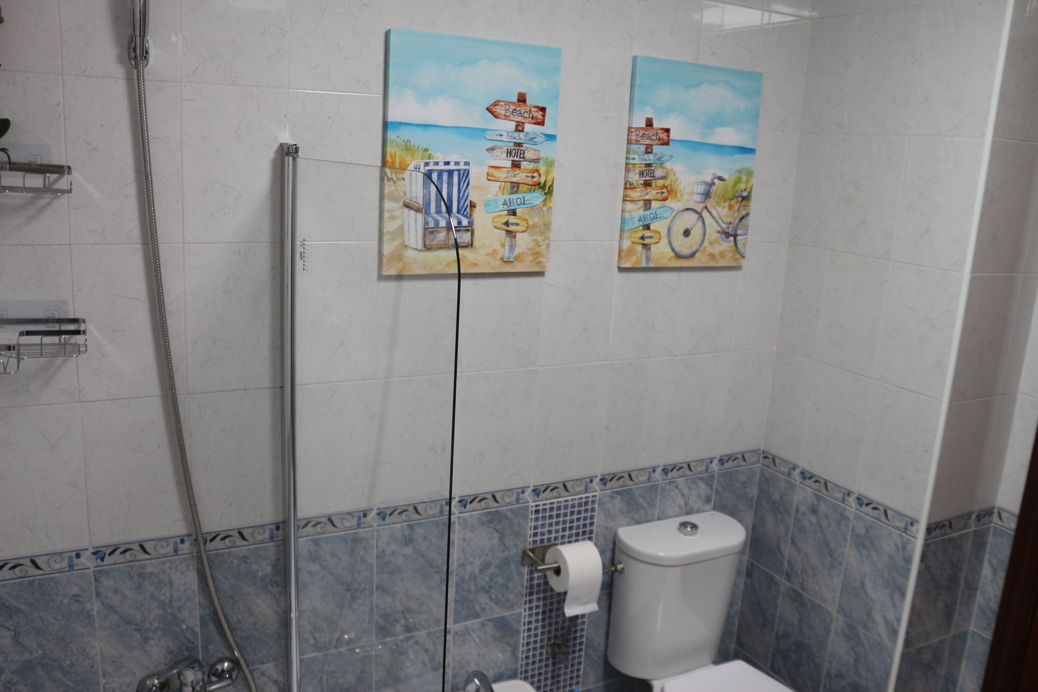 A bathroom with a shower, toilet, and two wall-mounted paintings depicting beach scenes.