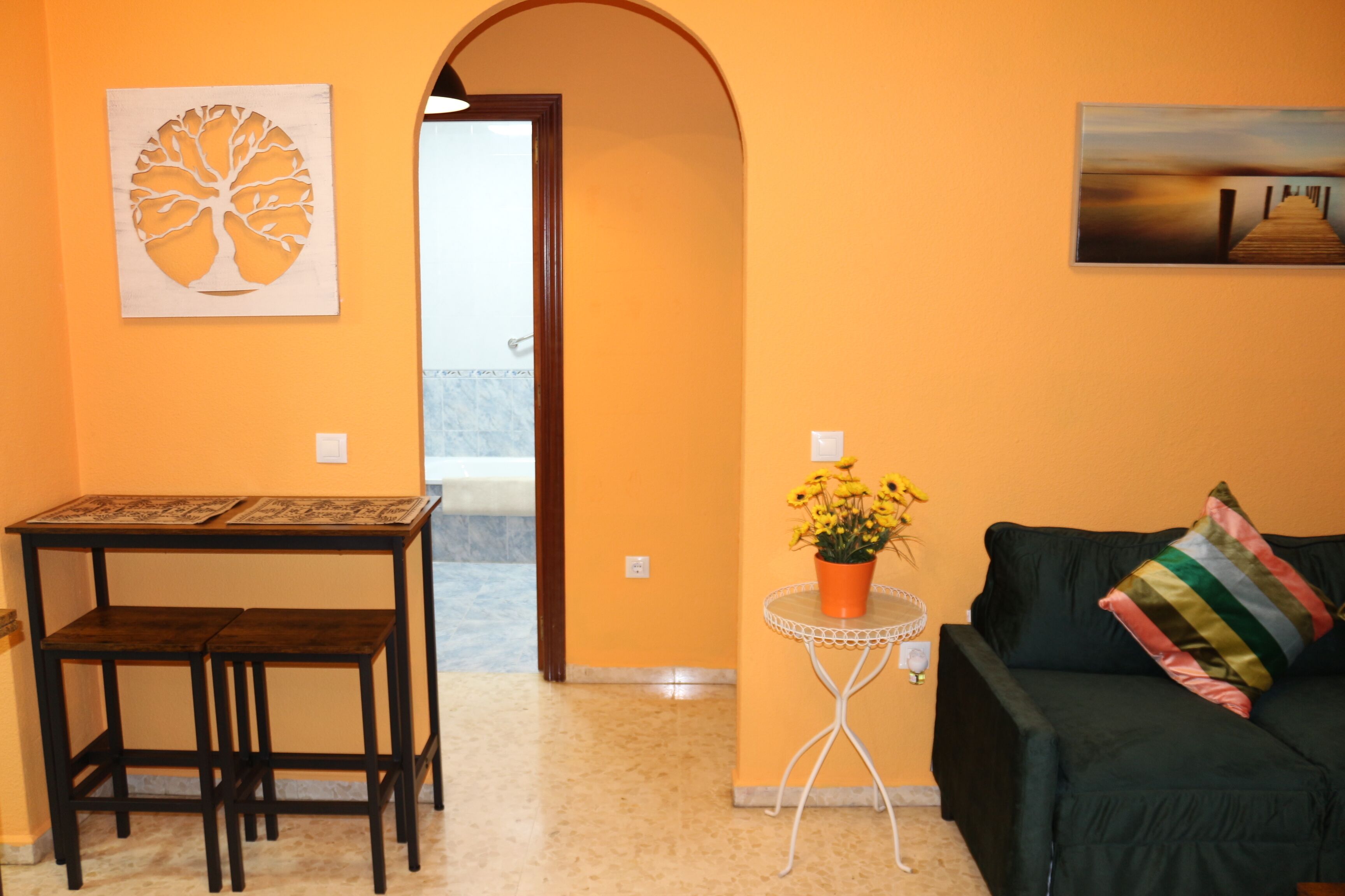 Una habitación con una mesa y sillas de madera, una pequeña mesa blanca con una planta en maceta, un sofá verde oscuro con un cojín a rayas y obras de arte en las paredes.