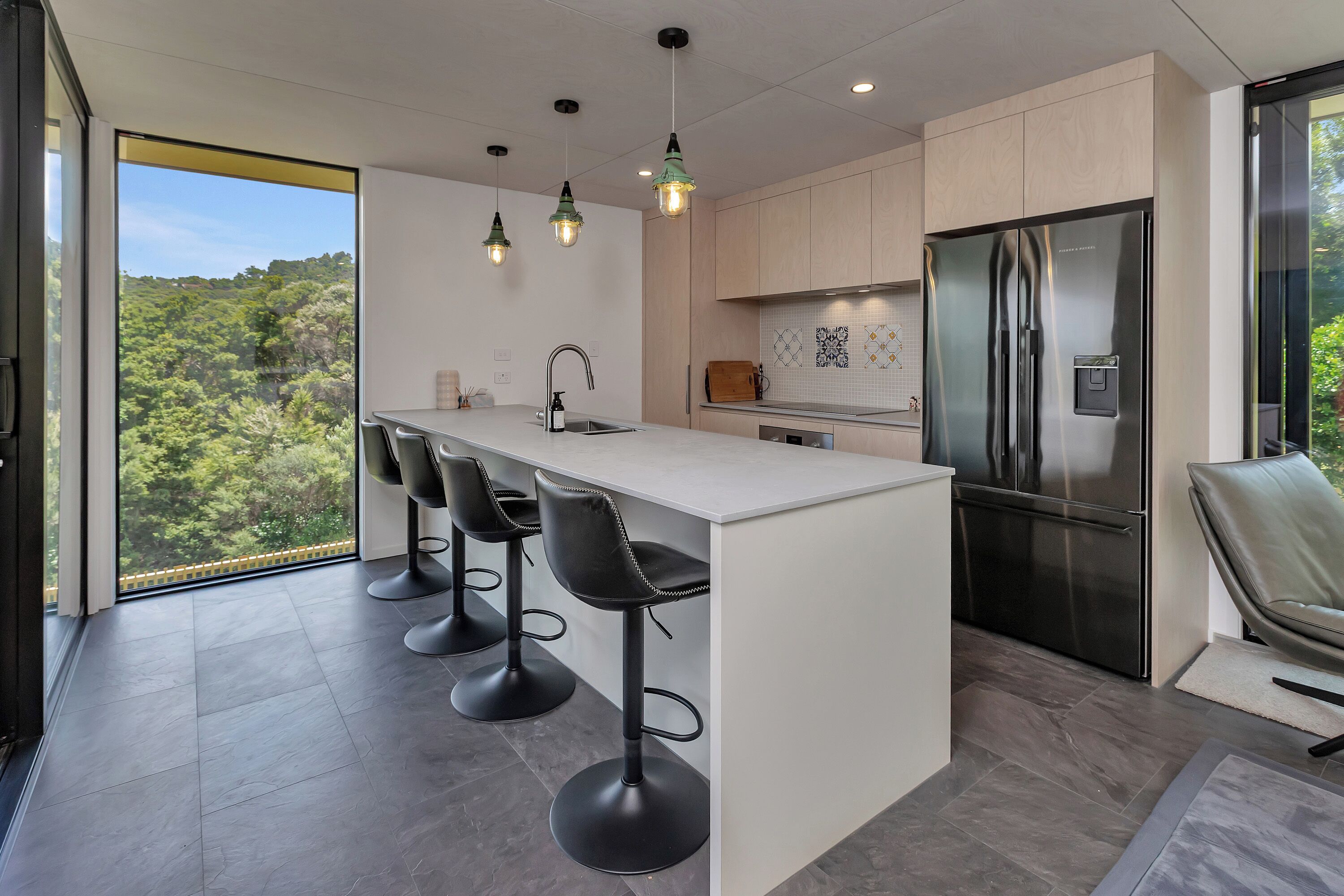 A modern kitchen with a central island, bar stools, a stainless steel refrigerator, and a view of greenery through large windows.