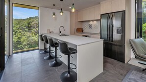 Cozinha moderna com ilha central, banquetas, geladeira de aço inoxidável e vista para o verde através de grandes janelas.