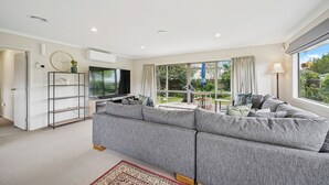 Ein modernes Wohnzimmer mit einem grauen Ecksofa, einem Flachbildfernseher und einem großen Fenster mit Blick auf den Garten.