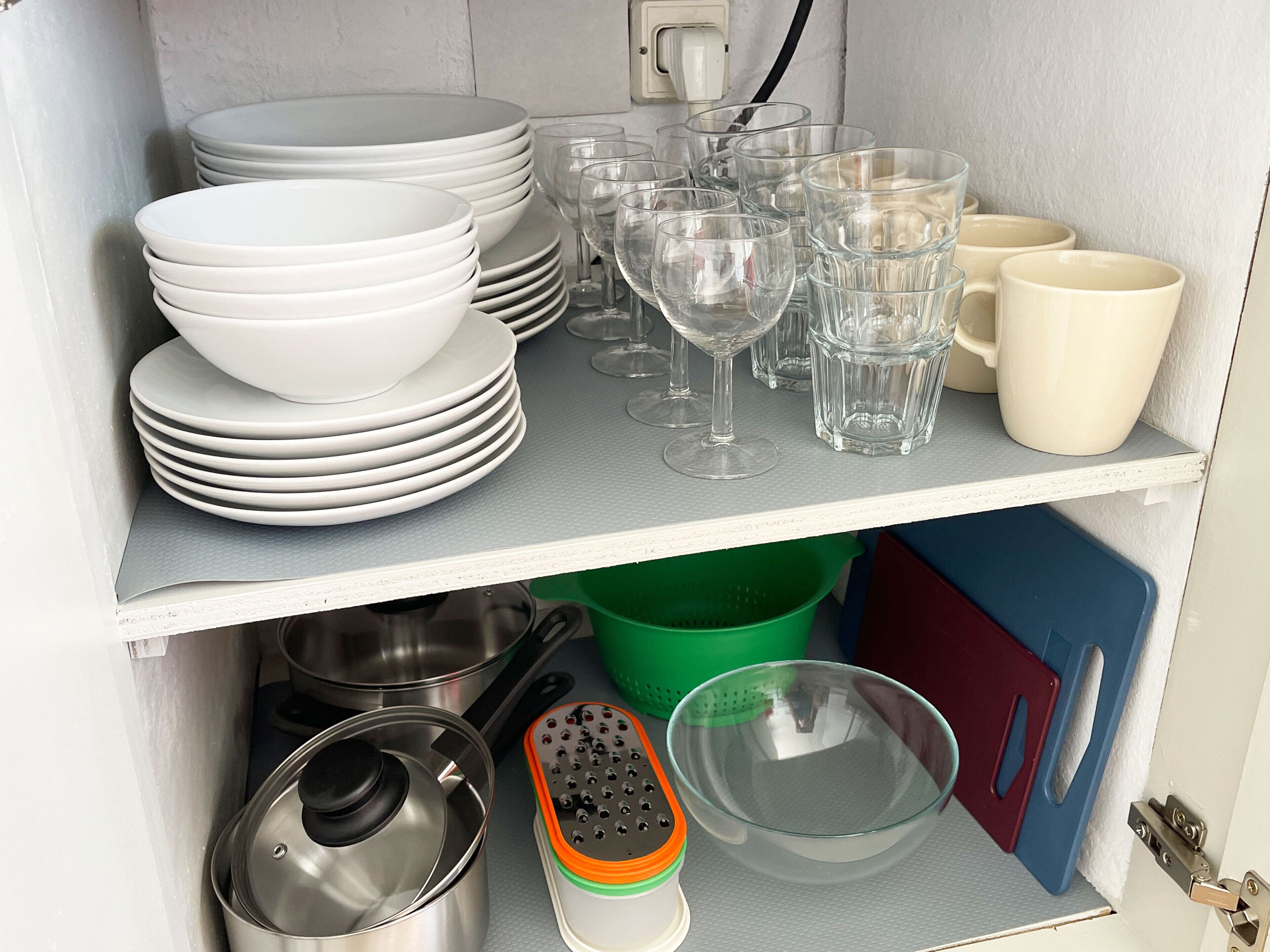 A kitchen cabinet with stacked white plates, clear drinking glasses, and a green plastic container.