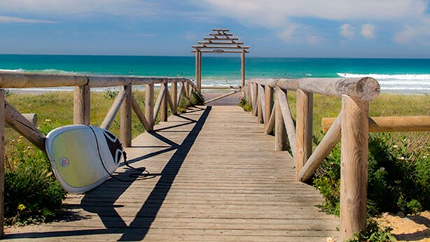 Una passerella di legno conduce a una spiaggia, con una tavola da surf appoggiata alla ringhiera.
