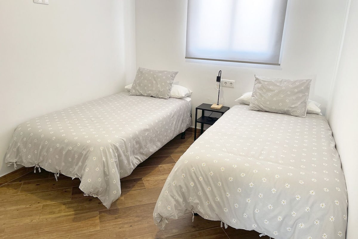A room with two single beds, each with a grey floral bedspread and matching pillows. There is a small bedside table with a lamp between the beds.