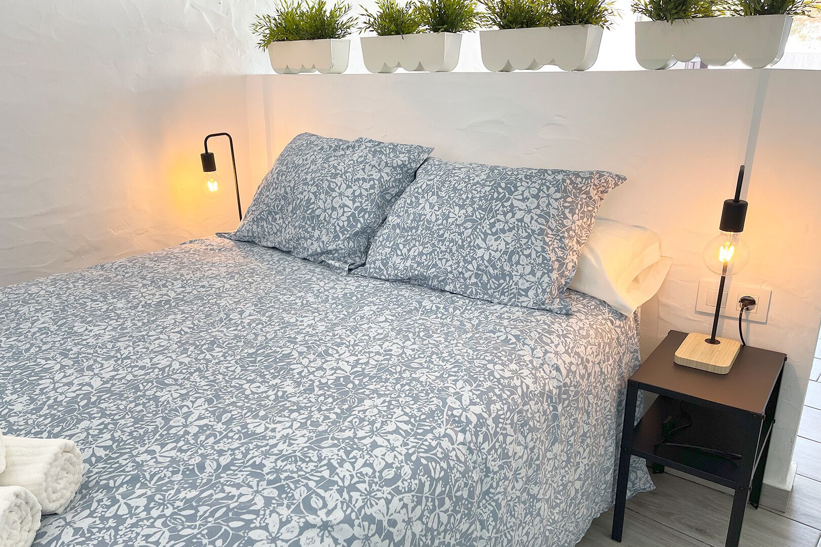 A neatly made bed with a floral-patterned bedspread, two pillows, and two bedside lamps. There are potted plants on a shelf above the bed.