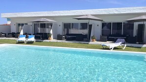 Zona piscina con sedie a sdraio e ombrelloni, piscina limpida di colore blu e un edificio con ampie finestre e posti a sedere all'aperto.