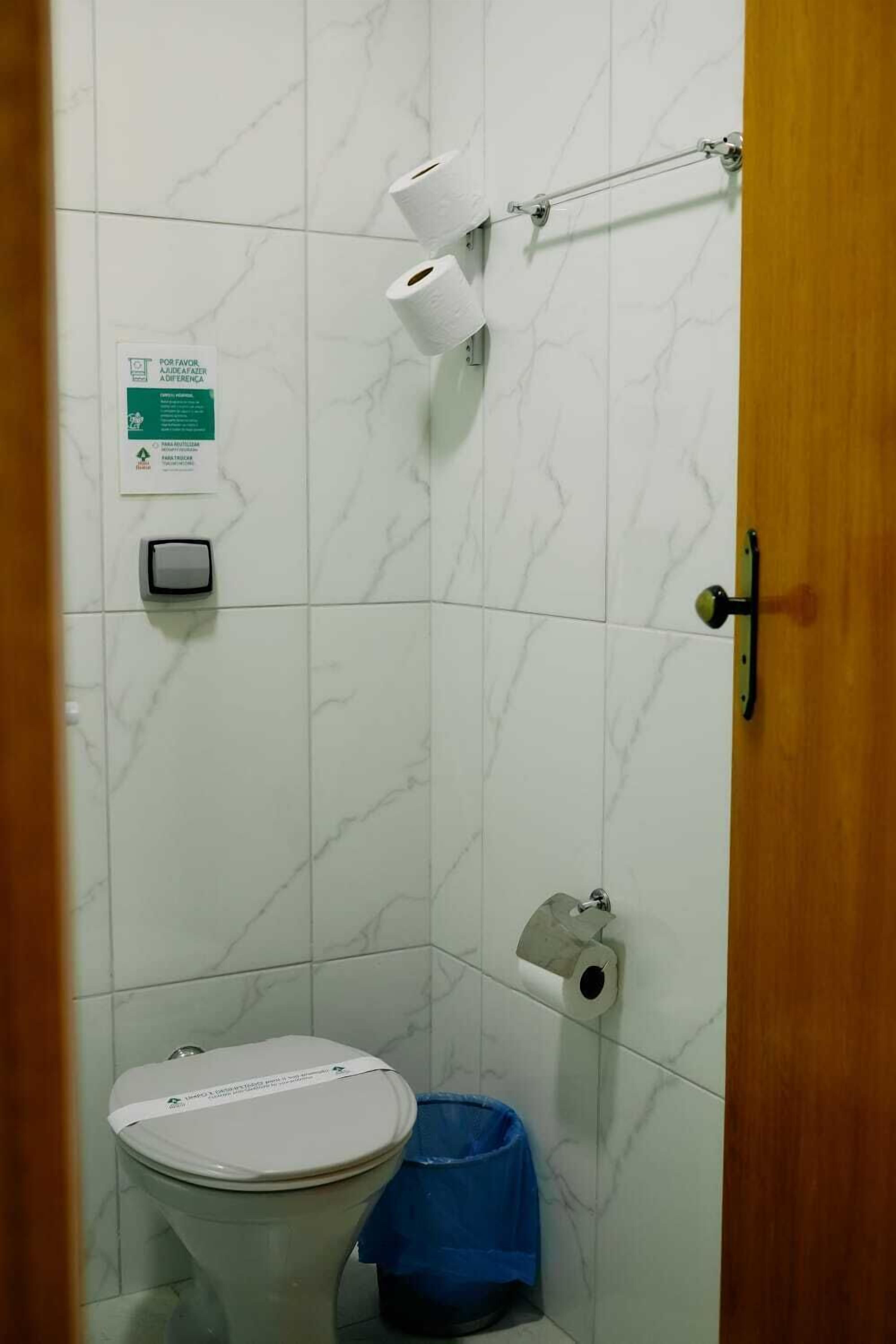 Un baño con inodoro, un bote de basura azul y dos rollos de papel higiénico en un perchero.