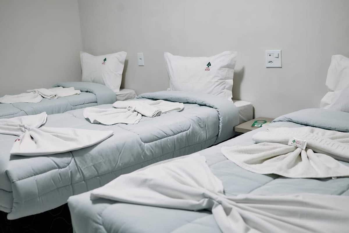 A hotel room with three beds, each with a white duvet and matching pillows. There are white towels on the beds, and a small table with a book and a green object.