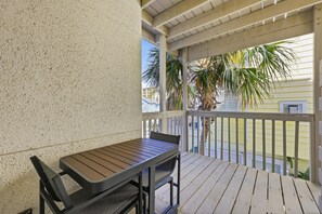 Uma área externa coberta com uma mesa e cadeiras de madeira, um corrimão branco e vista para um edifício amarelo e palmeiras.