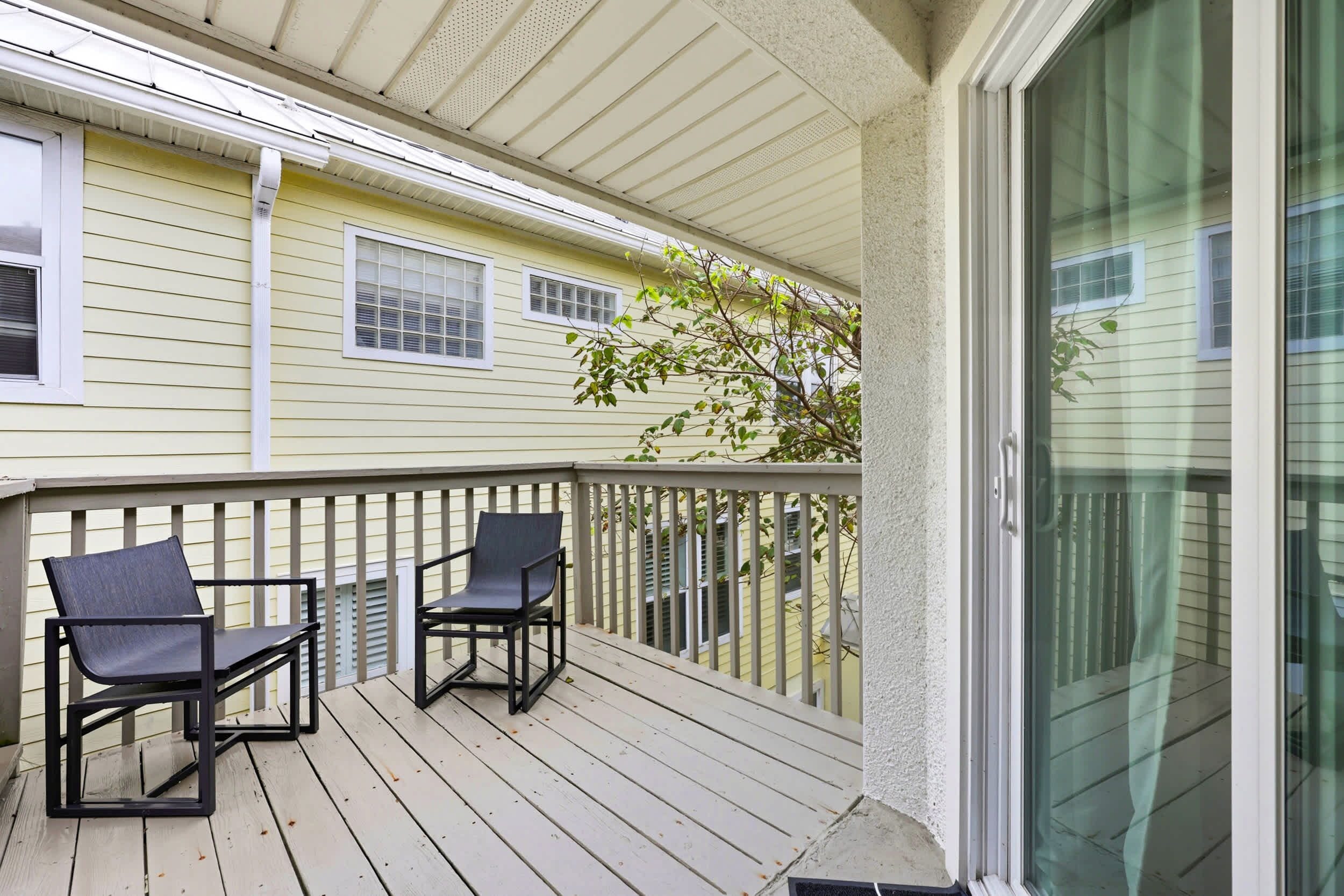 A covered outdoor balcony with two chairs and a glass door leading to a neighboring house.