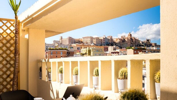 A balcony with a laptop, a cup, and a teapot, overlooking a cityscape with buildings and a palm tree. - LEOPOLDO ROOMS (Cagliari)
