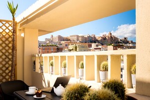 Un balcón con una computadora portátil, una taza y una tetera, con vistas a un paisaje urbano con edificios y una palmera.