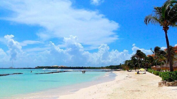 A beach with clear turquoise water, palm trees, and a few people in the distance. - Departamento cerca del mar 103 (Puerto Aventuras)