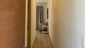 A hallway with a wooden door marked '104', a white wall, a ceiling with recessed lighting, and a glimpse of a room with a bed and curtains. - Departamento cerca del mar 103 (Puerto Aventuras)