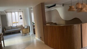 Un área de recepción moderna con un mostrador de madera curvada, una zona de estar con sofá y sillas, y lámparas colgantes tejidas.
