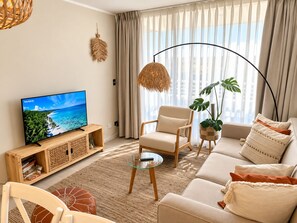 Un salon comprenant un téléviseur, un canapé, un fauteuil et une grande fenêtre avec des rideaux.