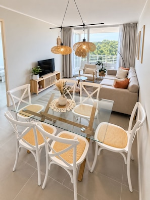 A modern living room with a glass dining table surrounded by white chairs with woven seats, a beige sofa, and a television on a wooden console. - El mejor lugar con Laguna paradisíaca (Las Cruces)