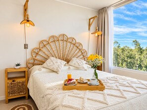 A bedroom with a large bed, a bedside table with a plant, a tray with breakfast items, and a view of trees through the window. - El mejor lugar con Laguna paradisíaca (Las Cruces)