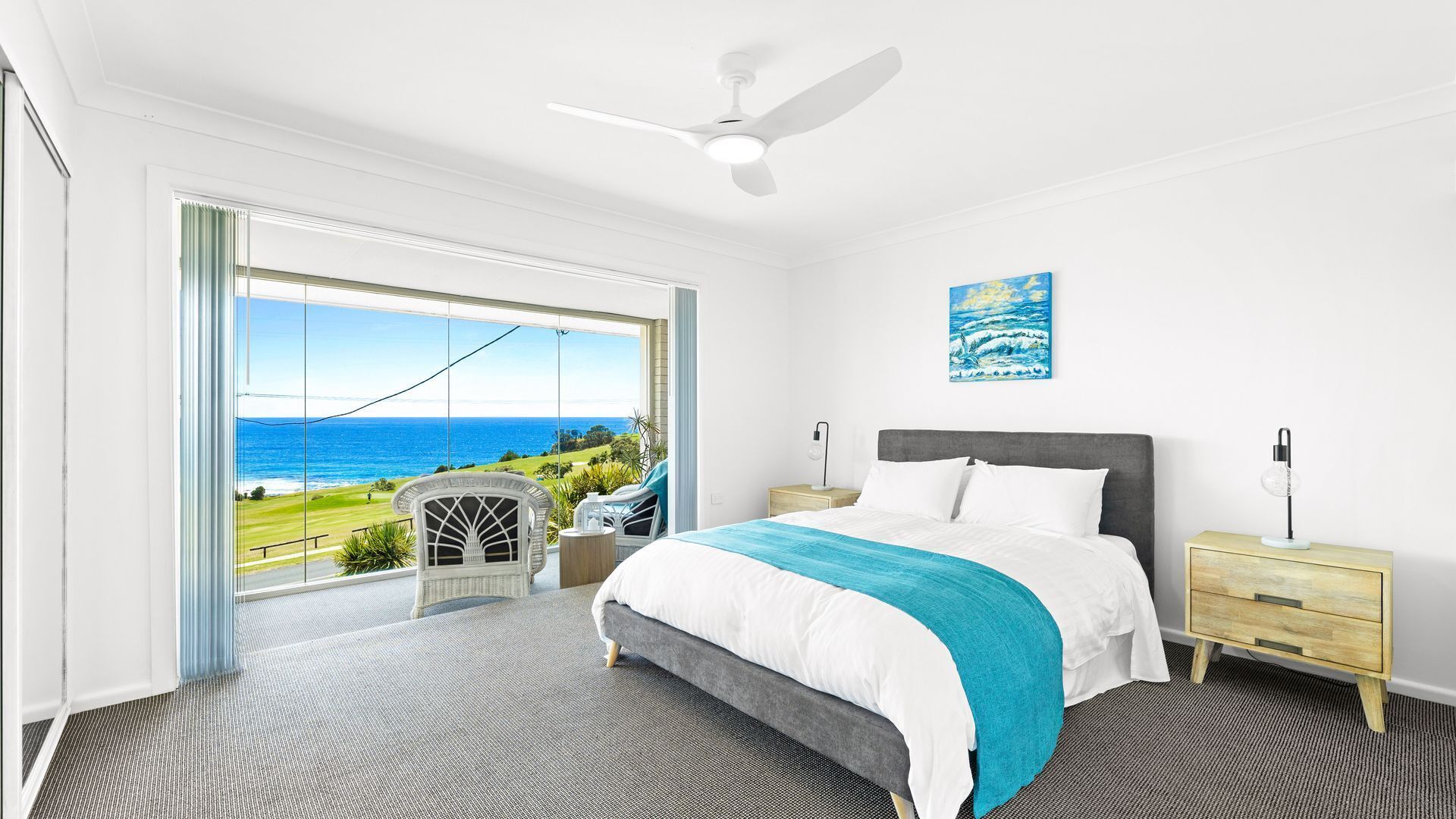 Quarto com cama, mesa de cabeceira, luminária e vista para o oceano.