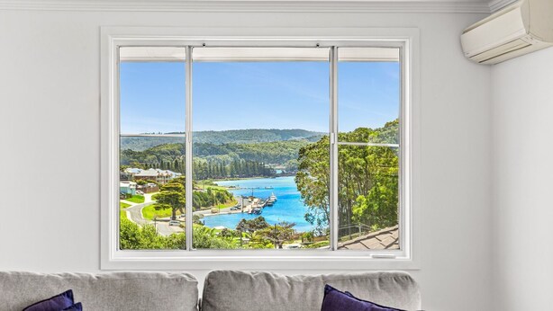 A room with a large window offering a view of a coastal area with a marina and lush greenery. - Golfers View Penthouse (Narooma)