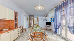 Zona de menjador amb una taula preparada per a quatre, envoltada de cadires. Hi ha una televisió a la paret, un llum al sostre i una finestra amb cortines.
