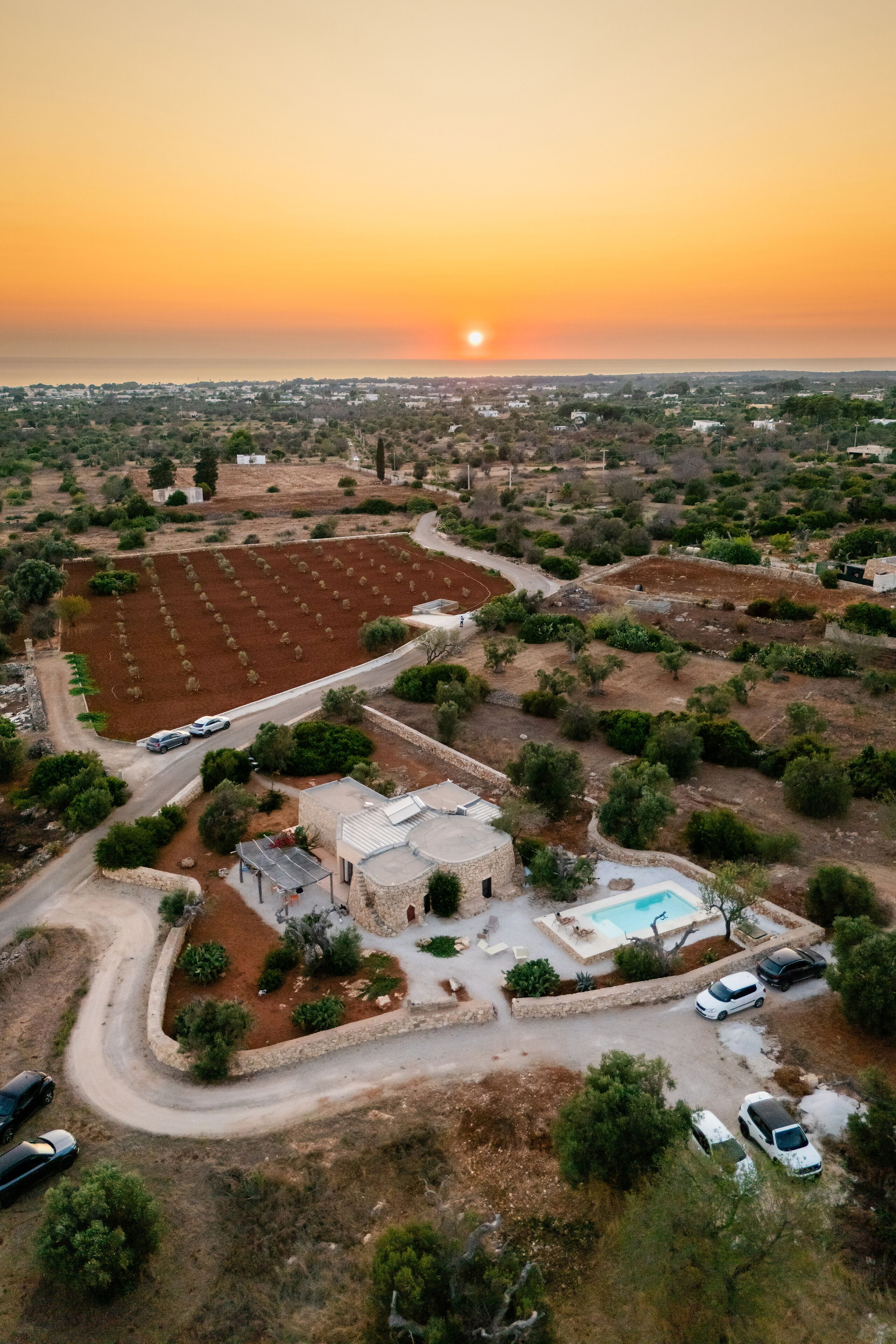 An aerial view of a property with a swimming pool, a small building, and a road with parked cars.