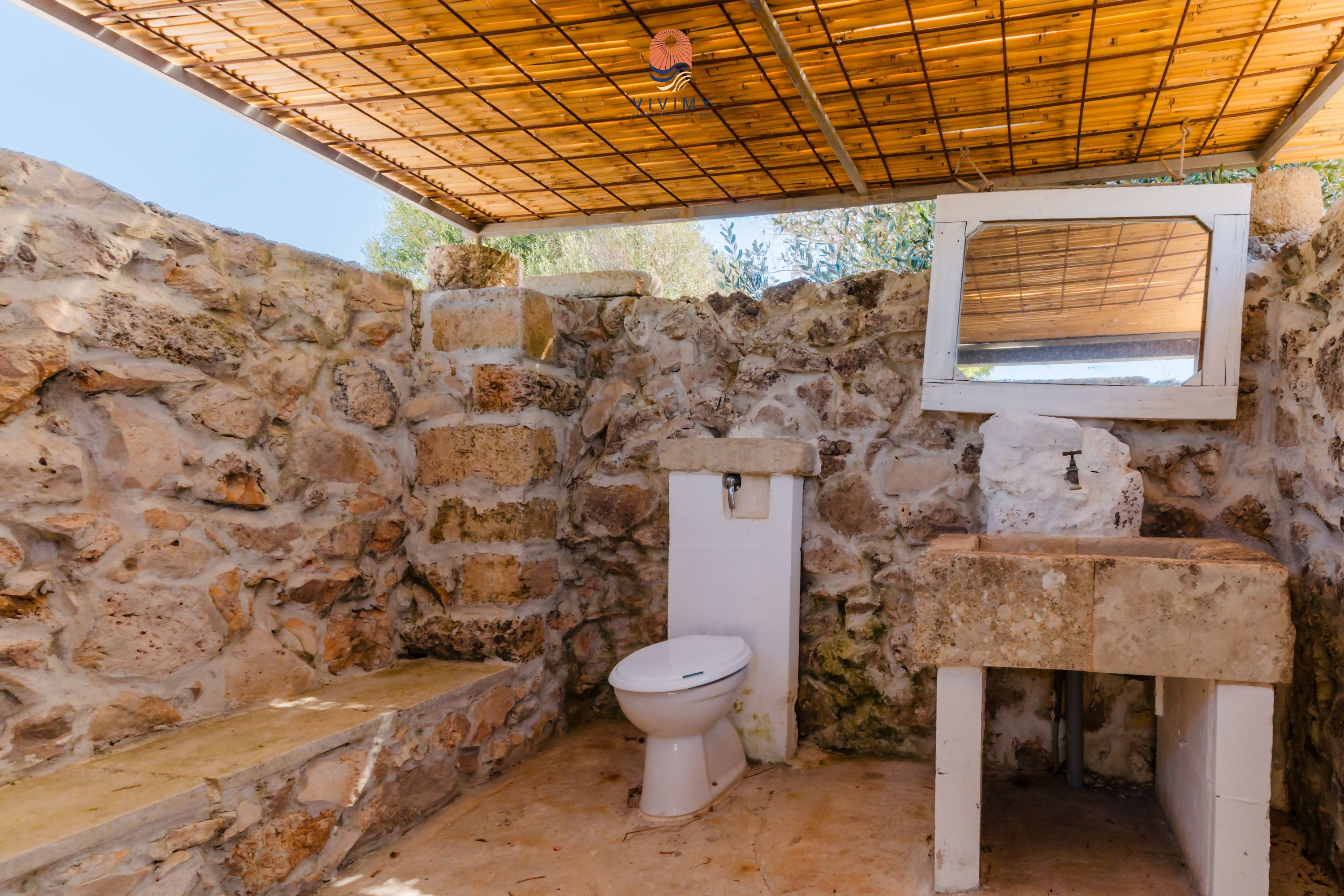 A rustic bathroom with a stone wall, a white toilet, and a wooden-framed sink.