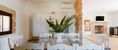 A dining area with a glass table and white chairs, a stone pillar, and a fireplace.