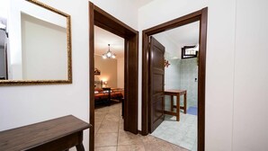 Un couloir avec une porte en bois qui donne sur une chambre à coucher, où se trouvent un lit et une table de chevet. Un miroir est accroché au mur et une petite table est placée dans un coin.