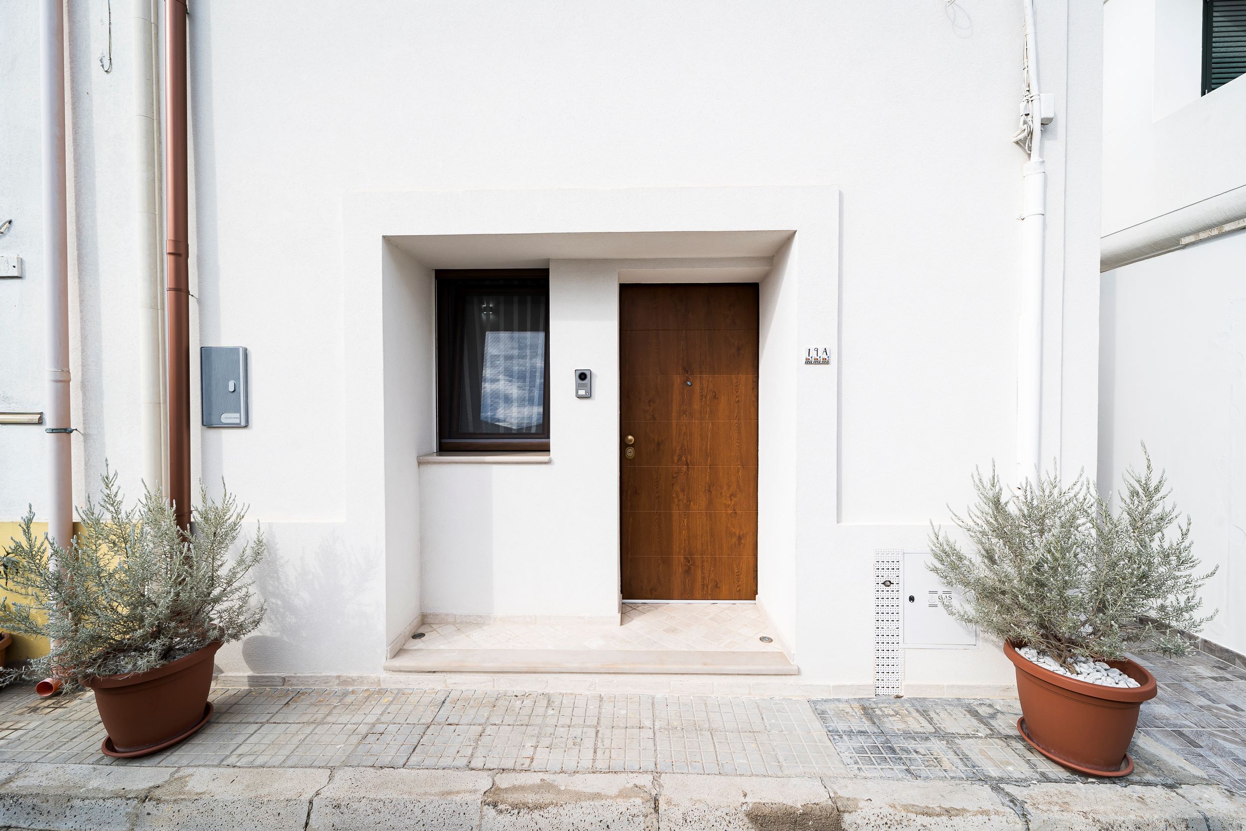 A white building with a wooden door and a window with a curtain. There are two potted plants on either side of the door.