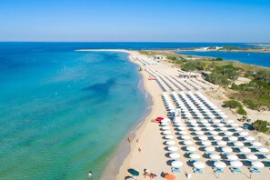 A beach with rows of sunbeds and umbrellas, a clear blue sea, and a few people walking along the shore. - Coraline Home by Vivimy (Felline)