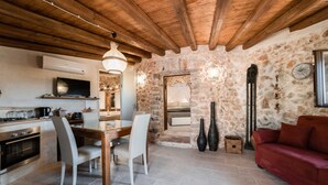 A rustic kitchen with a stone wall, wooden ceiling beams, a dining table with chairs, a red sofa, and a TV. - Trullo Puglia by Vivimy (Salve)