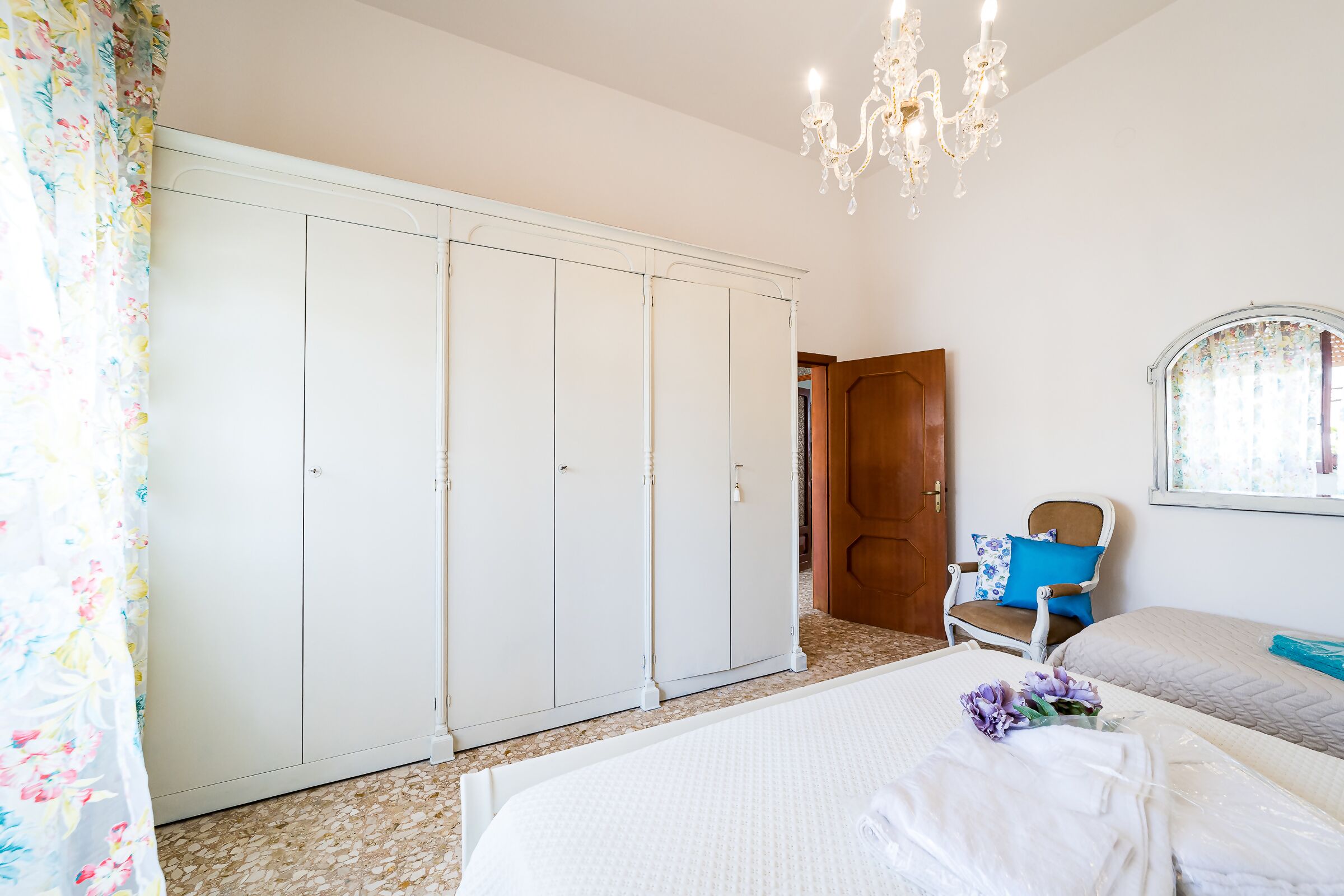 A bedroom with a large wardrobe, a bed with a white duvet, a chair with a blue cushion, and a chandelier.