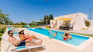 Zona de piscina con sillones de descanso, una mesa y un edificio de fachada blanca.