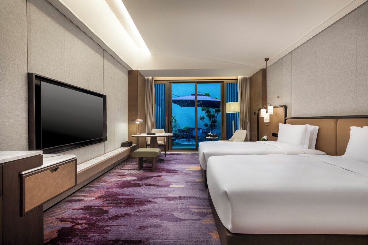 Ein Hotelzimmer mit großem Flachbildfernseher, zwei Betten, einer kleinen Sitzecke und Blick auf einen Außenpool.