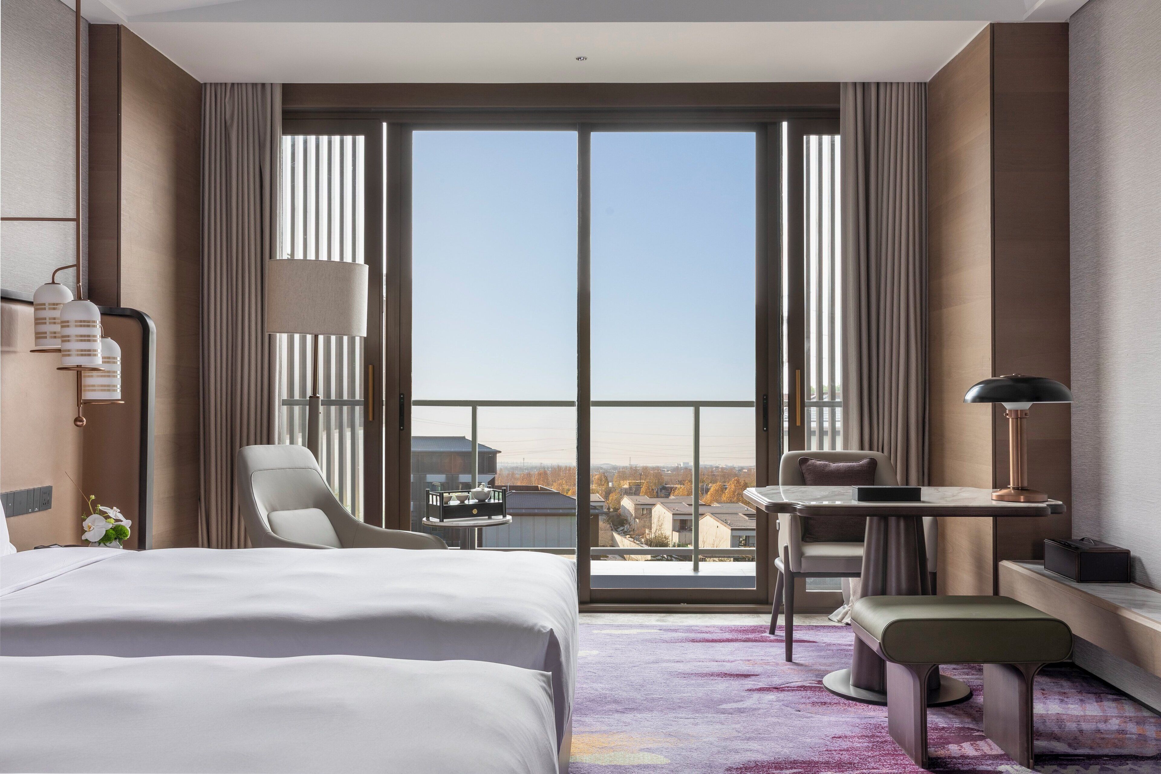 A hotel room with a large bed, a desk, a chair, and a view of the city through floor-to-ceiling windows.